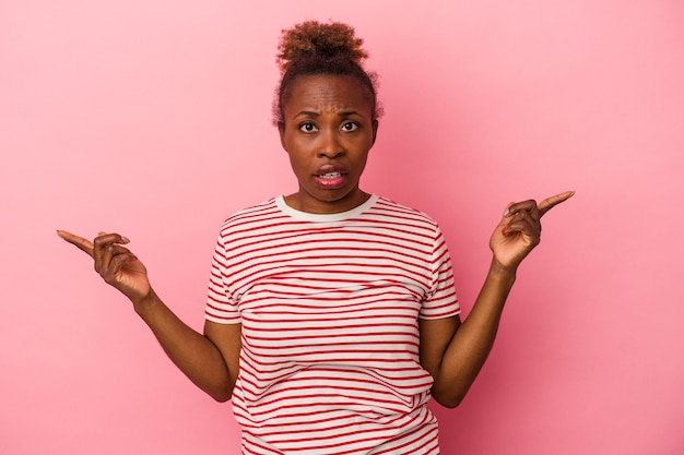 Jeune femme afro-américaine isolée sur fond rose pointant vers différents espaces de copie, en choisissant l'un d'entre eux, montrant avec le doigt.