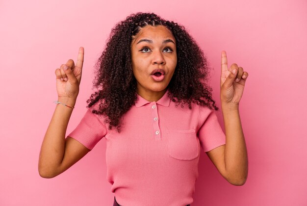 Jeune femme afro-américaine isolée sur fond rose pointant vers le haut avec la bouche ouverte.