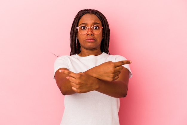 Une jeune femme afro-américaine isolée sur fond rose pointe sur le côté, essaie de choisir entre deux options.