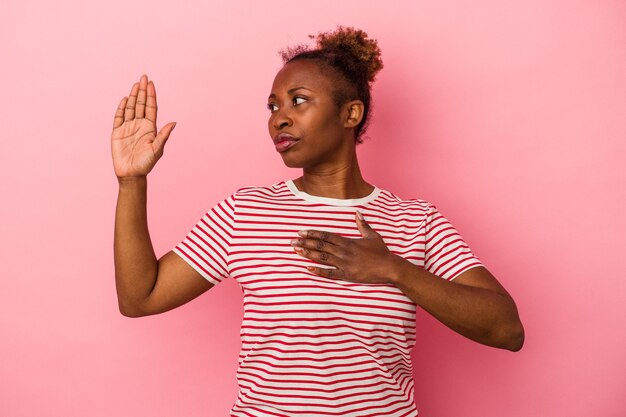 Jeune femme afro-américaine isolée sur fond rose prêtant serment, mettant la main sur la poitrine.