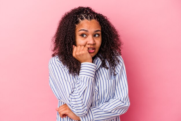 Jeune femme afro-américaine isolée sur fond rose se rongeant les ongles, nerveuse et très anxieuse.