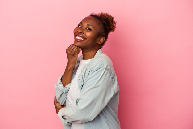 Jeune femme afro-américaine isolée sur fond rose souriante heureuse et confiante, touchant le menton avec la main.