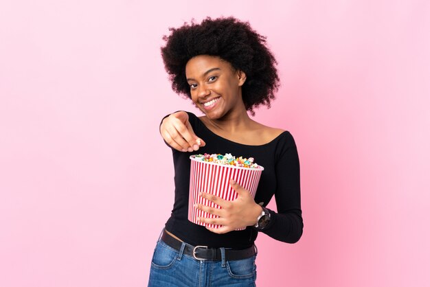 Jeune femme afro-américaine isolée sur fond rose tenant un gros seau de pop-corn tout en pointant vers l'avant