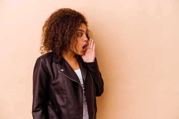 Jeune femme afro-américaine isolée sur un mur beige dit une nouvelle secrète de freinage à chaud et à côté