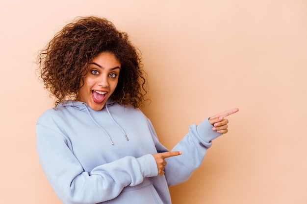 Jeune femme afro-américaine isolée sur un mur beige excité pointant avec l'index loin.