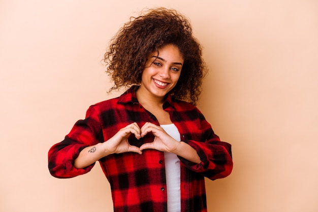 Jeune femme afro-américaine isolée sur un mur beige souriant et montrant une forme de coeur avec les mains.