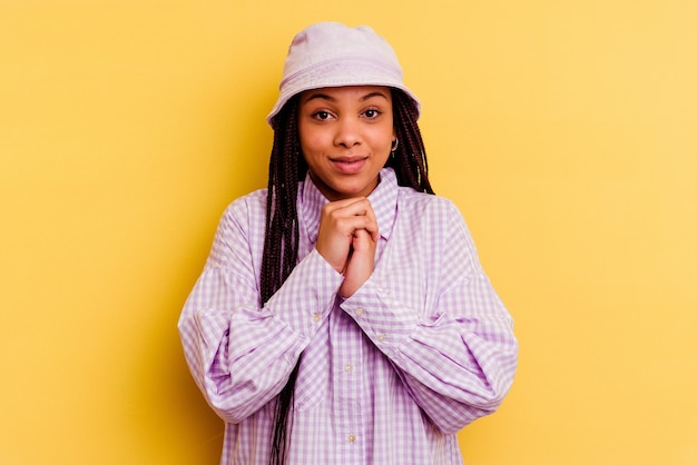 Jeune femme afro-américaine isolée sur le mur jaune garde les mains sous le menton, regarde joyeusement de côté.