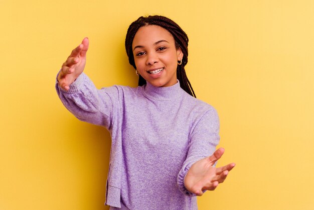 Jeune femme afro-américaine isolée sur le mur jaune se sent confiant en donnant un câlin à l'avant