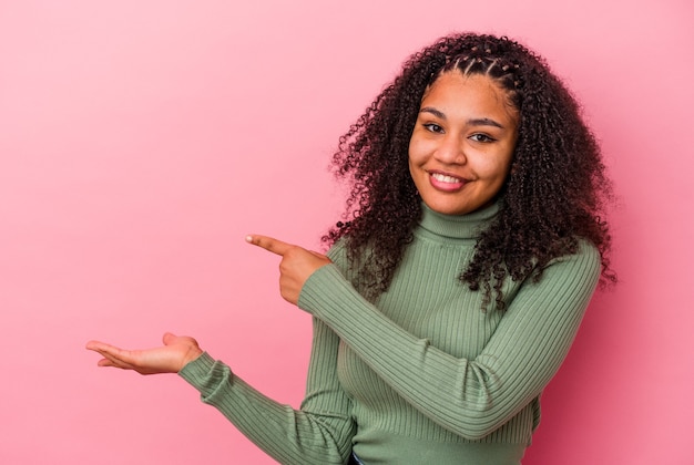 Jeune femme afro-américaine isolée sur un mur rose excité tenant un espace de copie sur la paume.