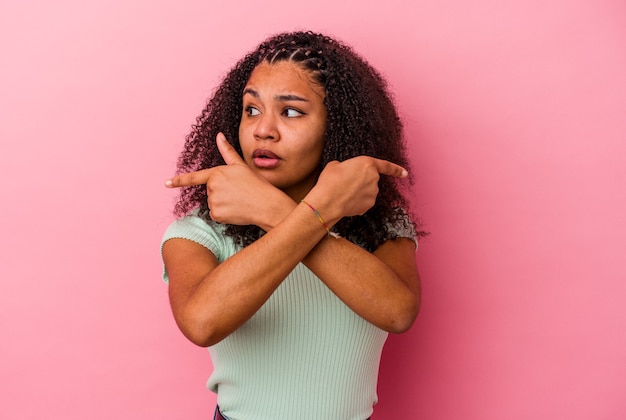 Jeune femme afro-américaine isolée sur le mur rose pointe sur le côté, essaie de choisir entre deux options