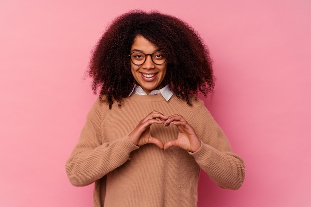 Jeune femme afro-américaine isolée sur mur rose souriant et montrant une forme de coeur avec les mains.