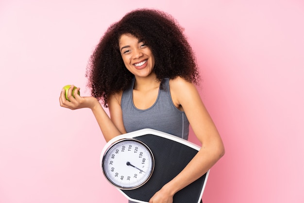 Jeune femme afro-américaine isolée sur rose avec peseuse et avec une pomme