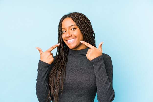 Jeune femme afro-américaine isolée sur des sourires de mur bleu, pointant les doigts sur la bouche.