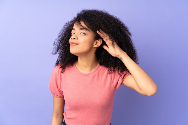 Jeune femme afro-américaine isolée sur violet écoutant quelque chose en mettant la main sur l'oreille