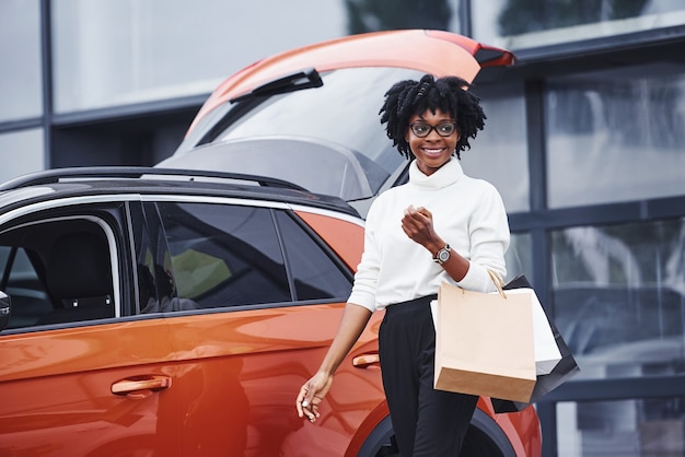 Une jeune femme afro-américaine à lunettes se tient à l'extérieur près d'une voiture moderne après avoir fait du shopping avec des colis.