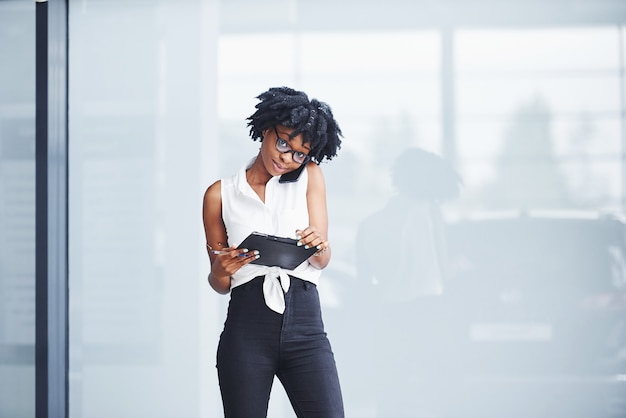 Jeune femme afro-américaine à lunettes se tient à l'intérieur et a un appel d'affaires.