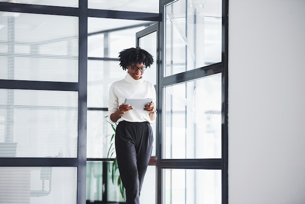 Une jeune femme afro-américaine à lunettes se tient à l'intérieur du bureau avec une tablette dans les mains.