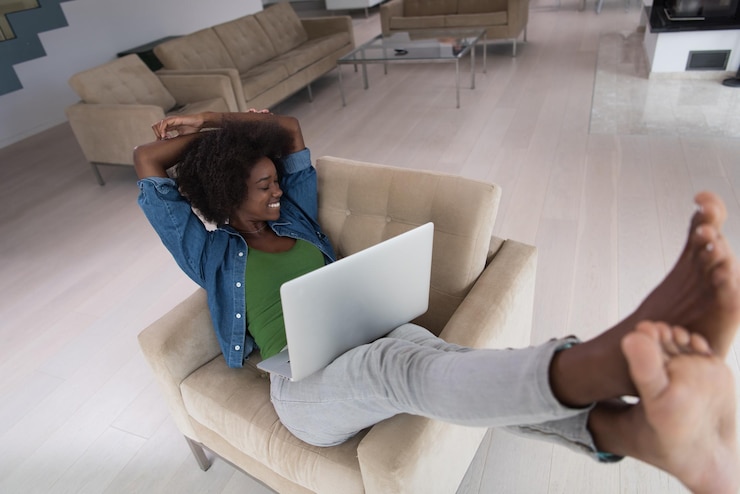 Photo jeune femme afro-américaine à la maison dans le fauteuil se relaxant dans son salon de luxe lisant un ordinateur portable surfer sur internet et travailler