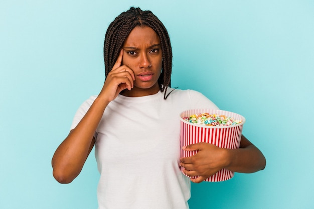 Jeune femme afro-américaine mangeant des pop corns isolés sur fond bleu pointant le temple avec le doigt, pensant, concentré sur une tâche.