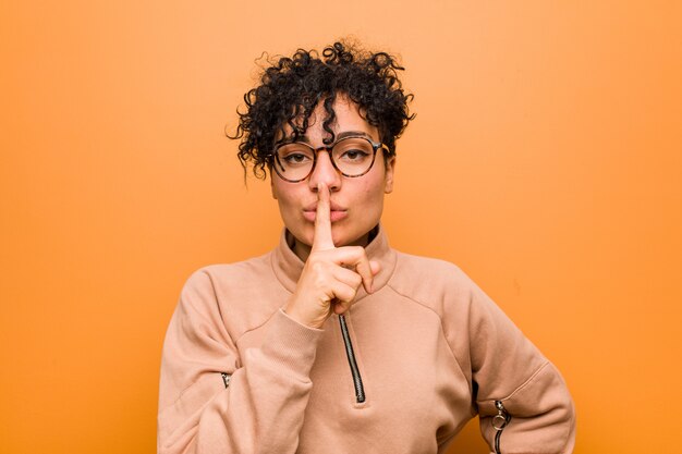 Jeune femme afro-américaine mixte contre un mur brun gardant un secret ou demandant le silence.