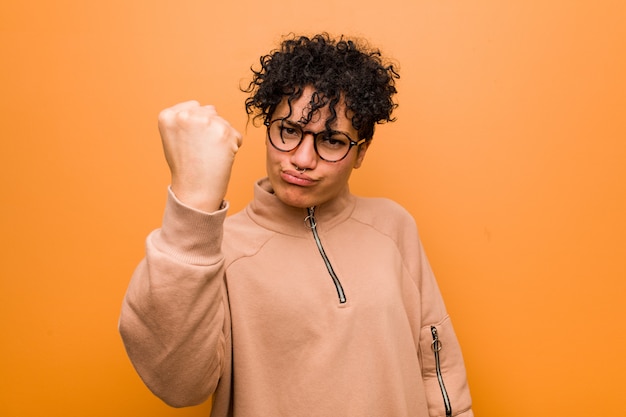 Jeune femme afro-américaine mixte contre un mur brun montrant le poing avec une expression faciale agressive.