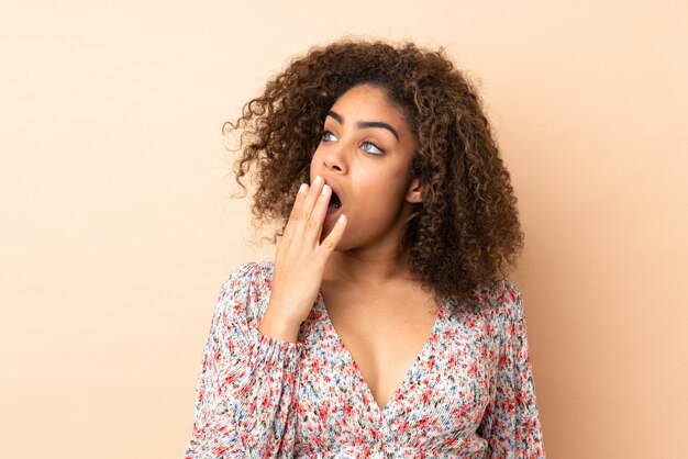 Jeune femme afro-américaine sur le mur beige bâillement et couvrant la bouche grande ouverte avec la main