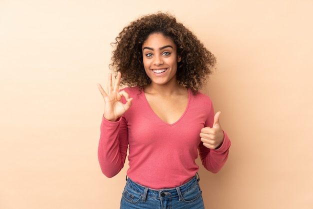 Jeune femme afro-américaine sur mur beige montrant le signe ok et le geste du pouce vers le haut