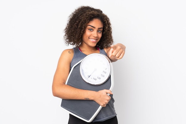 Jeune femme afro-américaine sur mur blanc tenant une machine de pesée et pointant vers l'avant