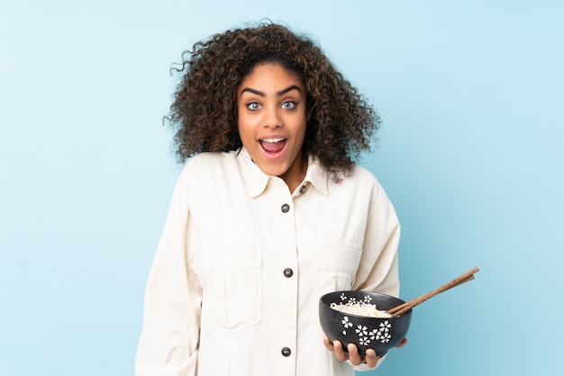 Jeune femme afro-américaine sur mur bleu avec une expression faciale surprise et choquée tout en tenant un bol de nouilles avec des baguettes