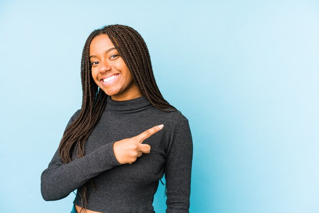 Jeune femme afro-américaine sur mur bleu souriant et pointant de côté, montrant quelque chose à l'espace vide.