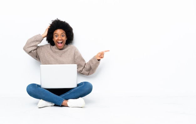 Jeune femme afro-américaine avec un ordinateur portable assis sur le sol surpris et pointant le doigt sur le côté