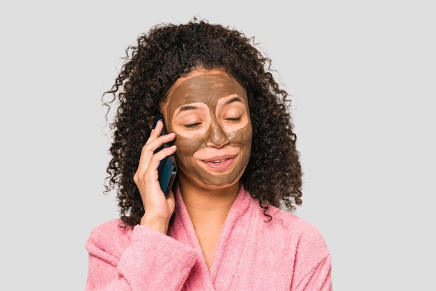 Jeune femme afro-américaine parlant au téléphone pendant qu'elle applique un masque facial isolé