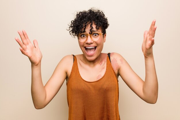 Jeune femme afro-américaine avec peau marque de naissance recevant une agréable surprise, excitée et levant les mains.