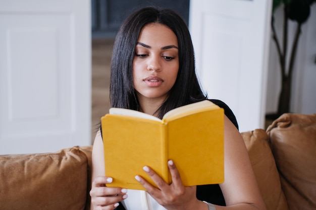 Photo jeune femme afro-américaine pensive assise sur un canapé en cuir lisant un livre à la maison