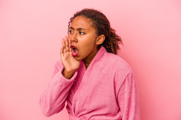 Jeune femme afro-américaine portant un peignoir isolé sur fond rose criant et tenant la paume près de la bouche ouverte.