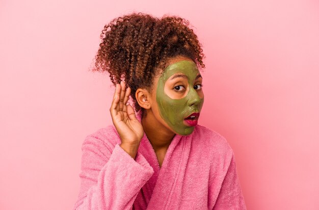 Jeune femme afro-américaine portant un peignoir et un masque facial isolé sur fond rose essayant d'écouter un potin.