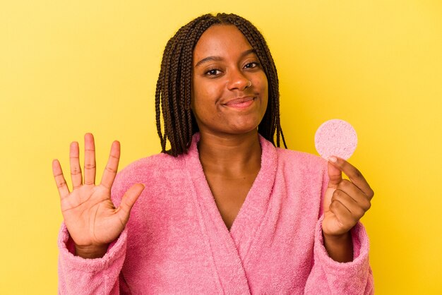 Jeune femme afro-américaine portant un peignoir tenant un disque de maquillage isolé sur fond jaune souriant joyeux montrant le numéro cinq avec les doigts.