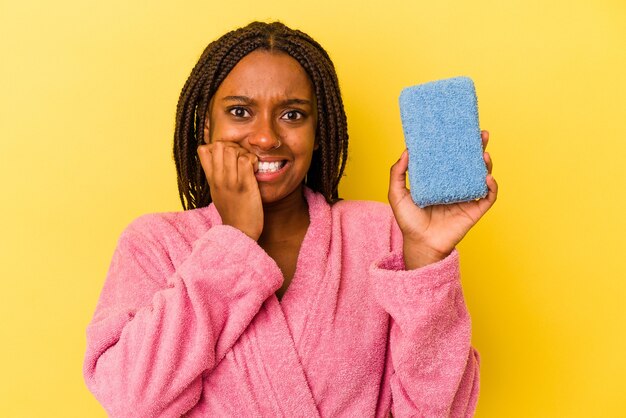 Jeune femme afro-américaine portant un peignoir tenant une éponge bleue isolée sur fond jaune se rongeant les ongles, nerveuse et très anxieuse.