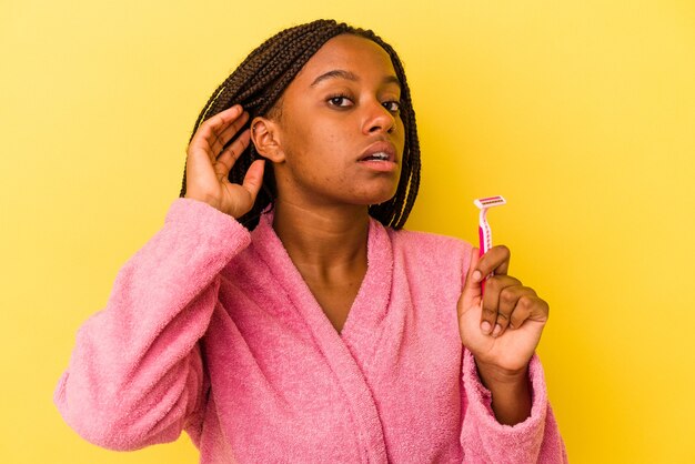 Jeune femme afro-américaine portant un peignoir tenant une lame de rasoir isolée sur fond jaune essayant d'écouter un potin.