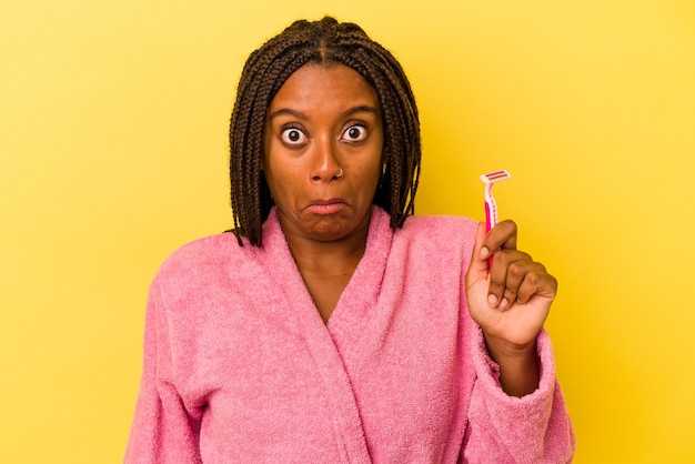 Jeune femme afro-américaine portant un peignoir tenant une lame de rasoir isolée sur fond jaune hausse les épaules et ouvre les yeux confus.