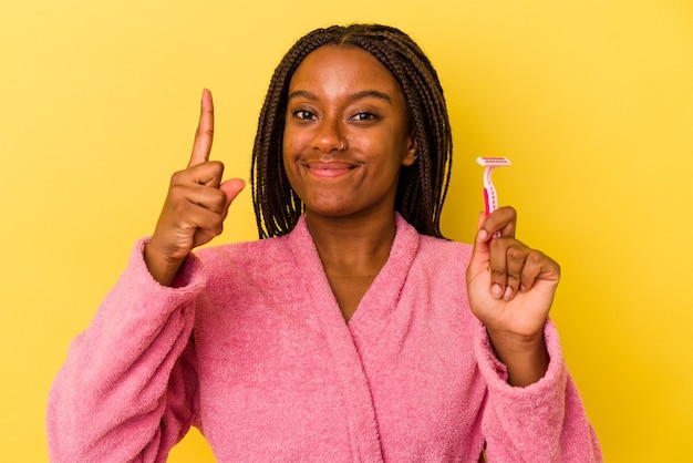 Jeune femme afro-américaine portant un peignoir tenant une lame de rasoir isolée sur fond jaune montrant le numéro un avec le doigt.