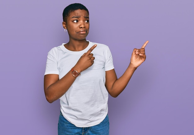 Jeune femme afro-américaine portant un t-shirt blanc décontracté pointant de côté inquiète et nerveuse des deux mains, expression inquiète et surprise