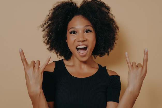 Jeune femme afro-américaine portant un t-shirt noir criant avec une expression faciale folle faisant un symbole rock-n-roll avec les mains comme une star de la musique, isolée sur fond de studio beige avec espace de copie