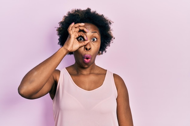 Jeune femme afro-américaine portant un t-shirt sans manches décontracté faisant un geste correct choqué par un œil de visage surpris regardant à travers les doigts expression incrédule