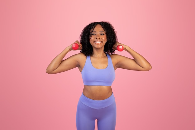 Jeune femme afro-américaine positive en vêtements de sport faisant de l'exercice pour les mains avec des haltères
