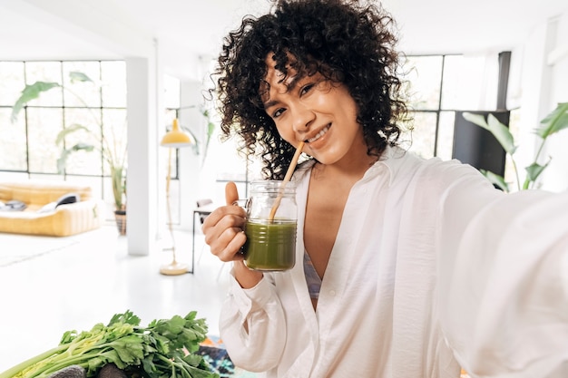 Jeune femme afro-américaine prenant un selfie boit du jus vert dans un loft lumineux Copy space