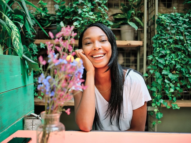 Jeune femme afro-américaine regardant la caméra et souriante assise dans un restaurant ou un café dans le jardin.