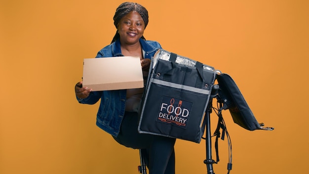 Jeune femme afro-américaine retirant une boîte de pizza du sac de livraison de nourriture pour un client dans le quartier local Courier travaillant sur demande livre efficacement de la restauration rapide avec un vélo comme moyen de transport