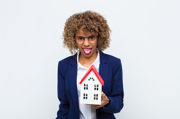 Jeune femme afro-américaine se sentant dégoûté et irrité, tirant la langue, détestant quelque chose de méchant et de dégoûtant avec un modèle de maison