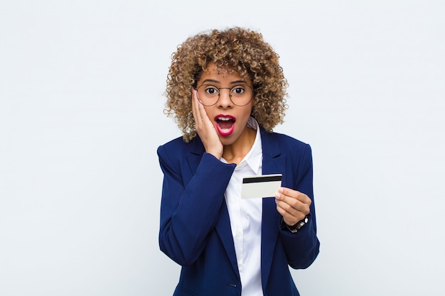 Jeune femme afro-américaine se sentir heureuse, excitée et surprise, regardant de côté avec les deux mains sur le visage avec une carte de crédit